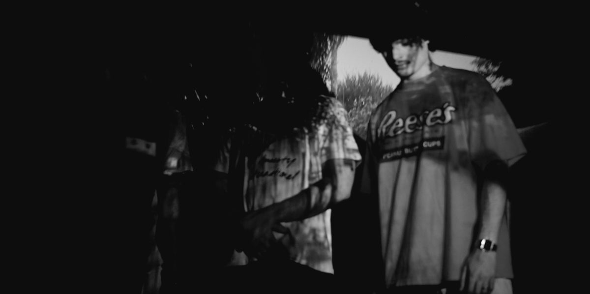 "Zwart-wit sfeerbeeld van drie jonge mannen in authentieke 90s vintage T-shirts, waaronder een oversized Reese's Peanut Butter Cups graphic tee, met een projectie van natuurbeelden op hun lichamen. Het beeld straalt een dromerige, rauwe en filmische sfeer uit, typerend voor subculturele streetwear en slow fashion. De jongens staan nonchalant in een schaduwrijke ruimte, wat de mysterieuze en artistieke esthetiek van het merk versterkt. Ideaal voor liefhebbers van nostalgische branding, Amerikaanse popcultuur