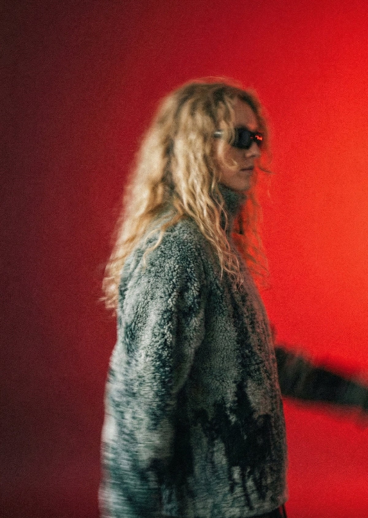 Model met zonnebril en grijsgroene teddyjas tegen een rood verlichte achtergrond bij Slow Fashion Vintage.