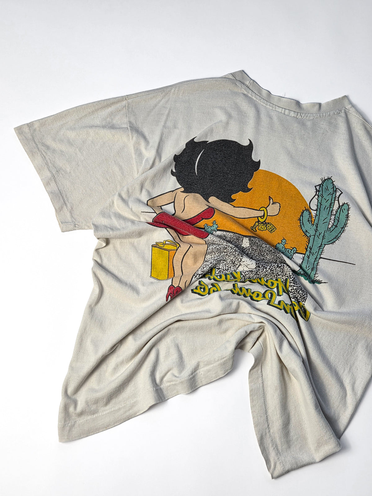 Achterkant van een ecru Betty Boop vintage T-shirt uit de jaren '90 met grote faded Route 66 backprint van Betty Boop in een Amerikaans woestijnlandschap, neergelegd en verkreukeld.