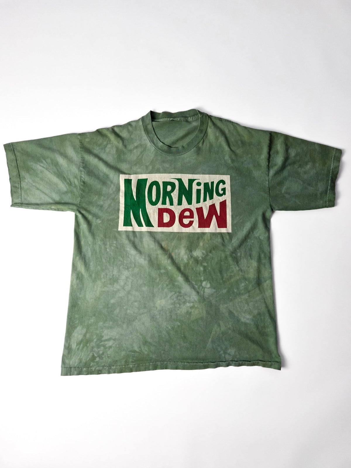 Groen Grateful Dead Morning Dew T-shirt met rood-groene logoprint op een gebroken wit vlak, volledig plat gefotografeerd.