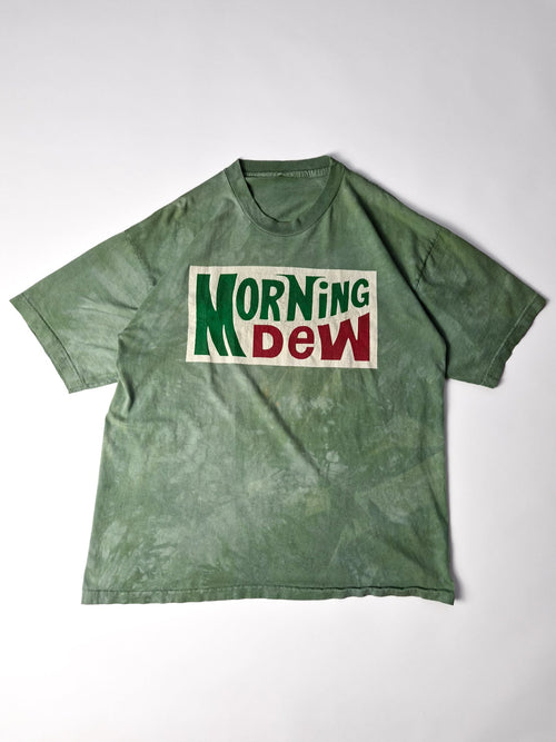 Groen Grateful Dead Morning Dew T-shirt met rood-groene logoprint op een gebroken wit vlak, volledig plat gefotografeerd.