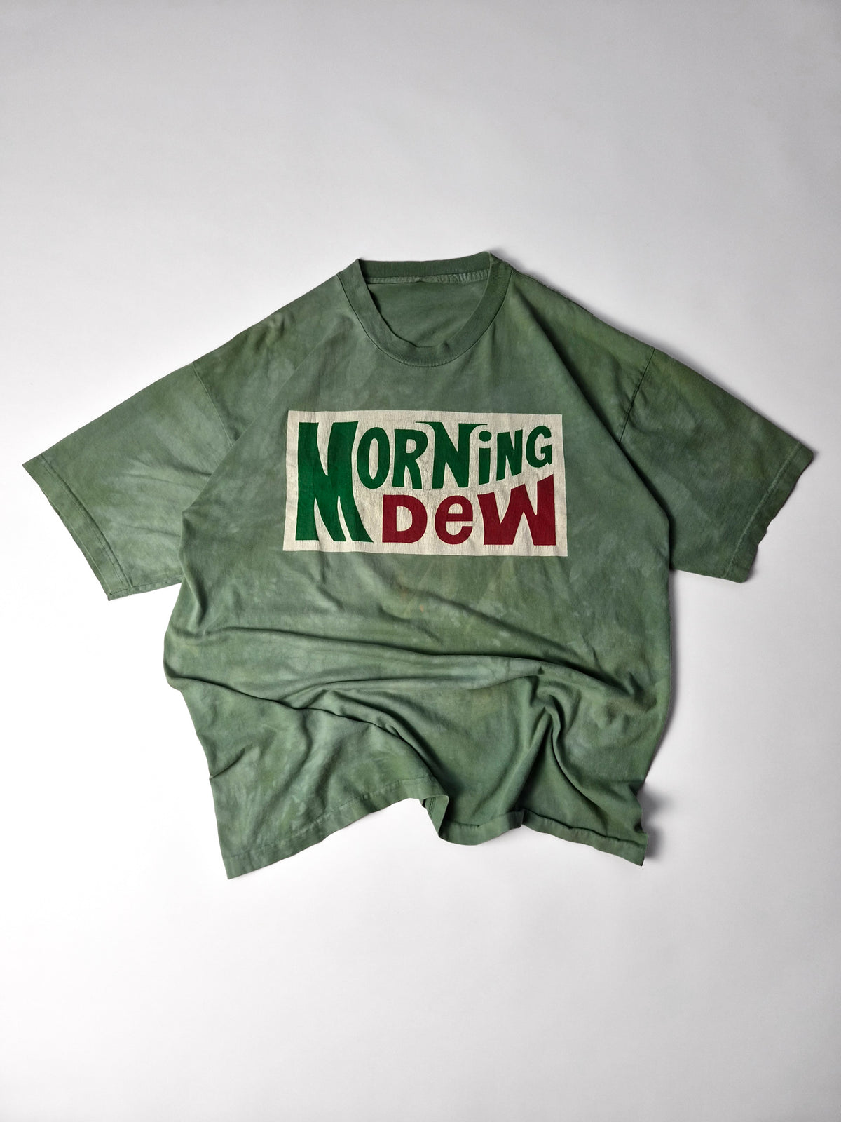 Groen Grateful Dead Morning Dew T-shirt met rood-groene logoprint op een gebroken wit vlak, volledig plat gefotografeerd.
