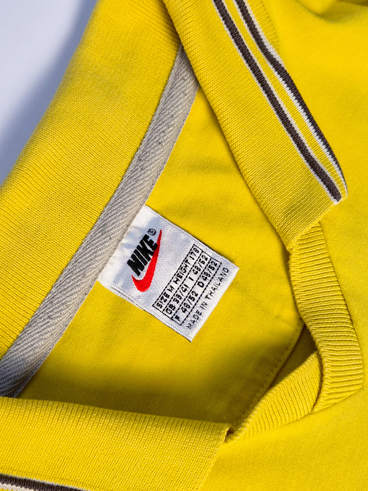 Nike label in geel vintage rugby shirt uit de jaren 90, maat M en Made in Thailand, met rode Swoosh en gestreepte kraag.