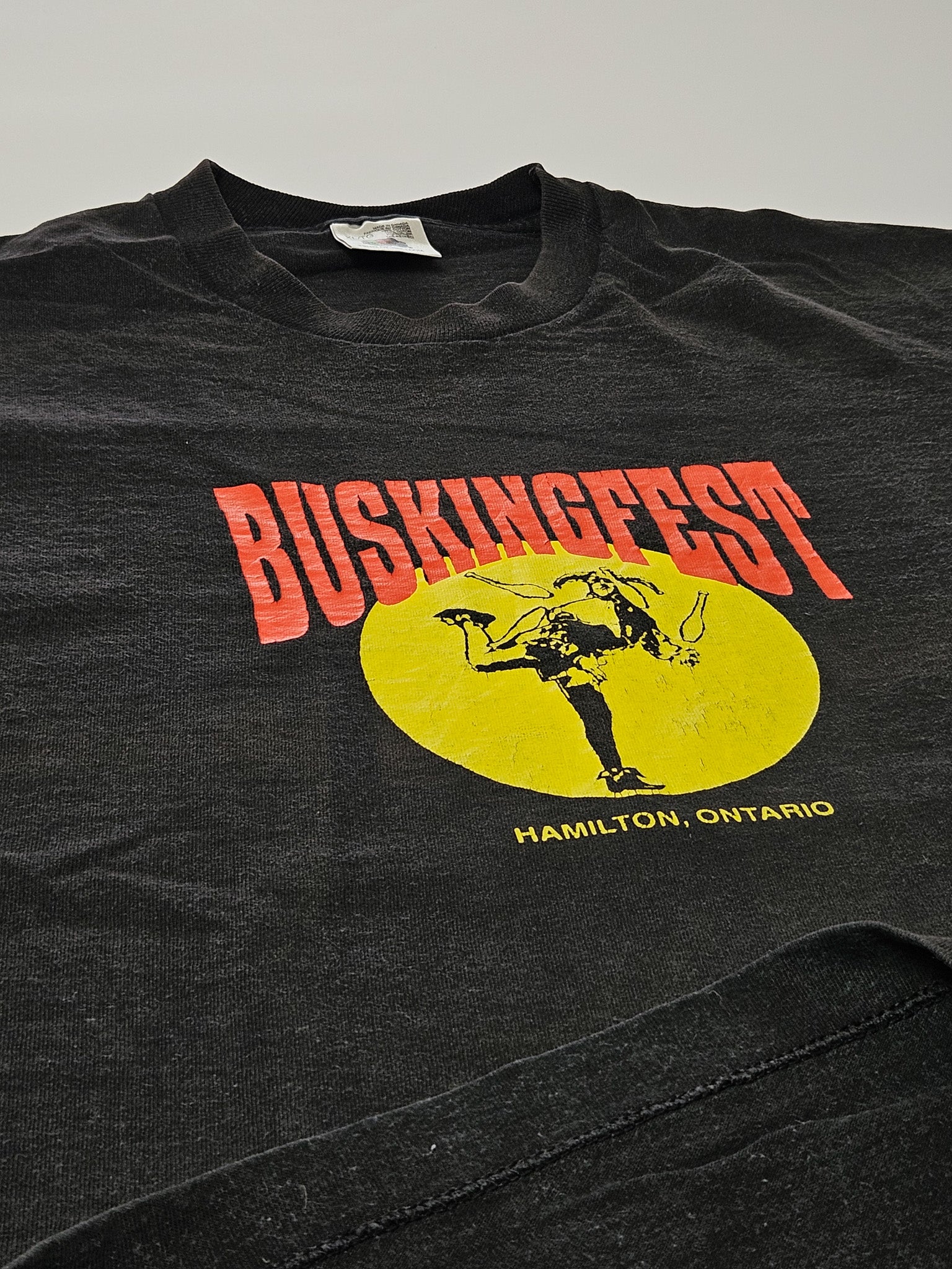1990s vintage BuskingFest T-shirt in zwart, met grote geel-rode print van straatartiest en tekst ‘Hamilton, Ontario’ op de voorkant