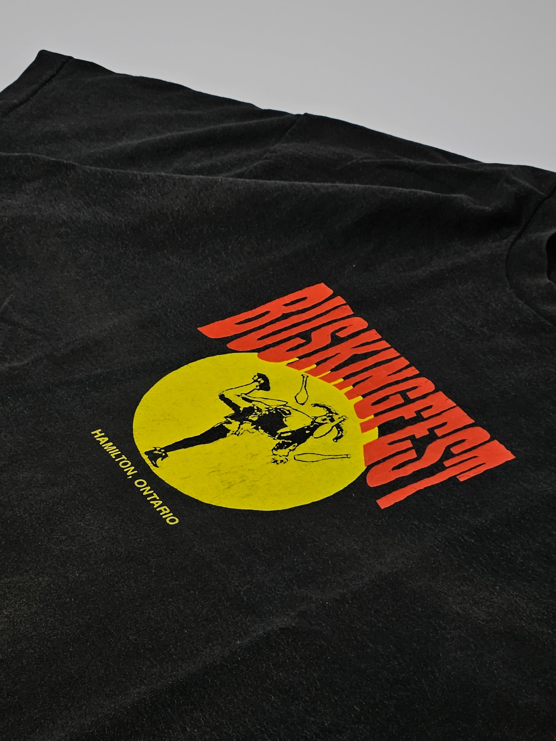 1990s vintage BuskingFest T-shirt in zwart, met grote geel-rode print van straatartiest en tekst ‘Hamilton, Ontario’ op de voorkant