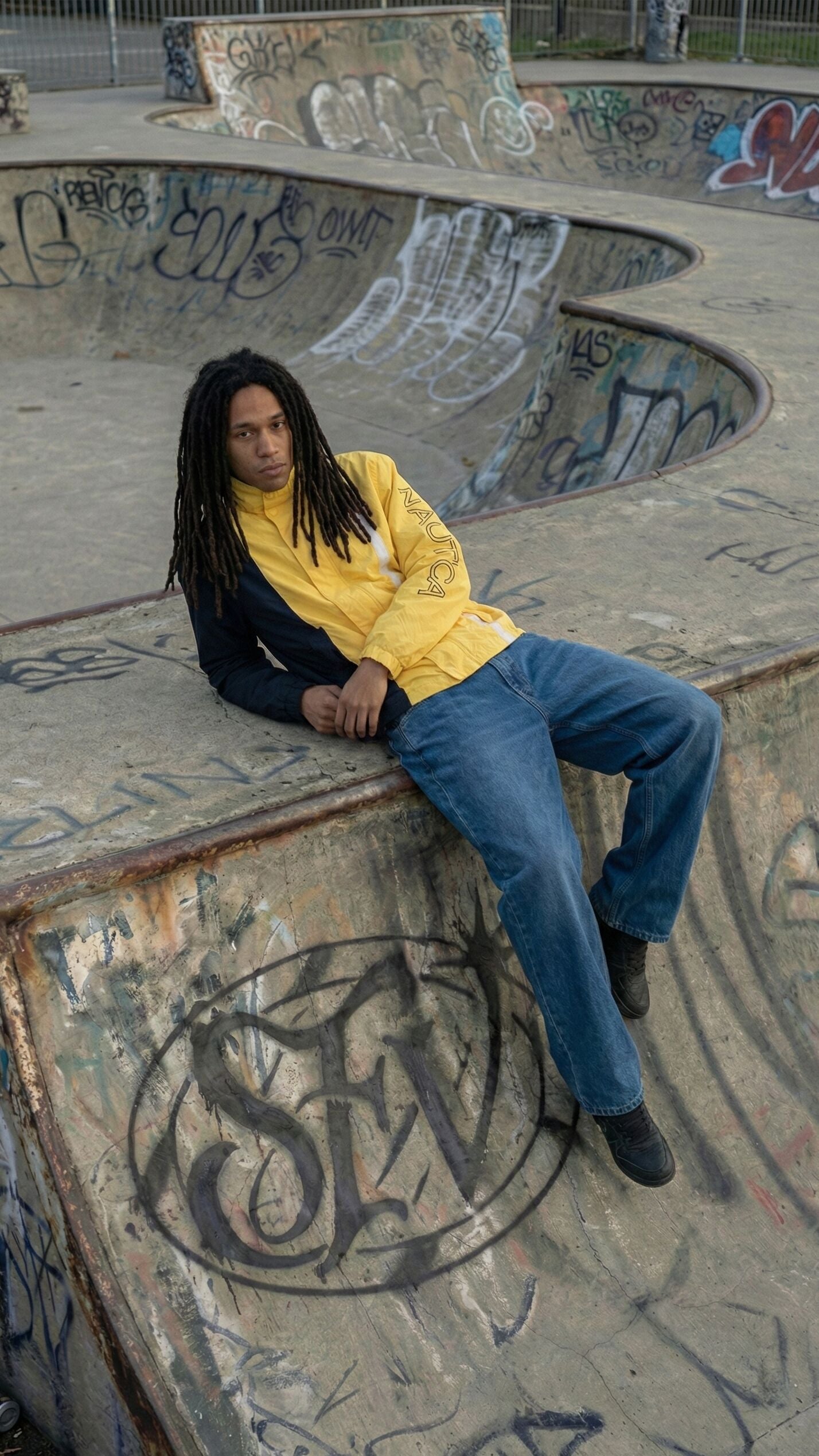 Jonge man in een geel-zwarte vintage windbreakerjack, ontspannen liggend tegen een verweerde betonnen skatepark-ramp met graffiti. Lange dreadlocks, casual jeans, urban setting met spuitbussen en ruwe streetstyle sfeer, passend bij authentieke 90s streetwear van Slow Fashion Vintage.