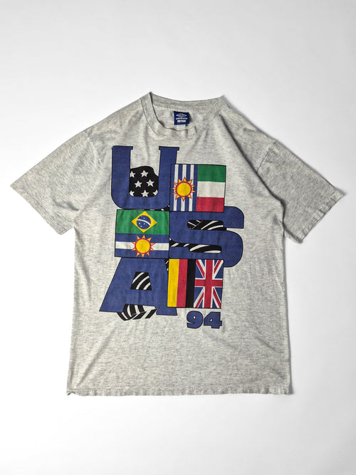 Lichtgrijs vintage Umbro T-shirt uit de jaren ’90 met grote blauwe USA ’94-letters en een kleurrijke vlaggenprint op de voorkant.