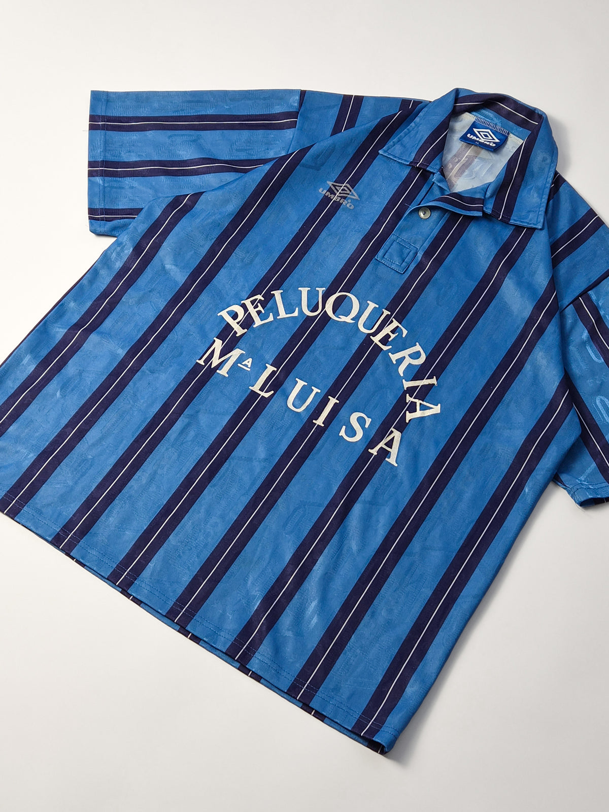Vintage Umbro voetbalshirt uit de jaren '90 in blauw en navy met Peluquería Maluisa sponsor, voorkant flatlay, schuin gefotografeerd.